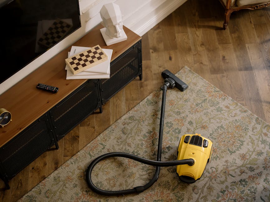 A yellow vacuum cleaner with a long black hose and metal wand attachment lies on a patterned area rug in a living room, positioned near a wooden media console with a stack of white papers, a chessboard, a small white bust sculpture, and a remote control. The room features a hardwood floor, white skirting boards, and a wall with a mounted flat-screen television. Natural light illuminates the space, emphasizing the clean and well-maintained surfaces. Carpet Cleaning Islington specializes in surface cleaning, deep cleaning, and sanitisation for residential spaces, ensuring carpets and floors are spotless and hygienic.