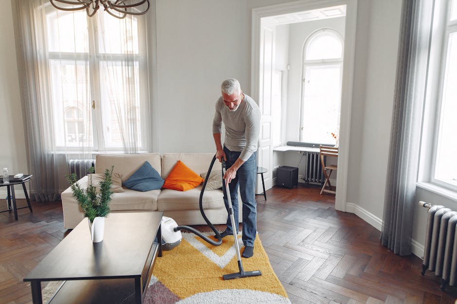 A person kneeling on a floral patterned carpet in a residential setting, using a yellow portable vacuum cleaner with a black hose and a digital display on top. The individual's hands rest on the vacuum's control panel as they operate it, with the surrounding area showing a wooden floor and soft lighting that highlights the cleanliness of the carpet. This scene exemplifies deep cleaning or surface cleaning, reflecting professional or domestic carpet maintenance, coordinated by Carpet Cleaning Islington, a specialist company in the cleaning services category.