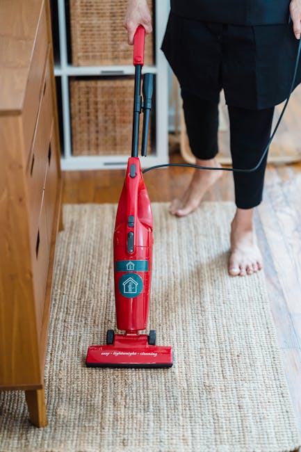 A man with white hair and a beard, wearing a light gray long-sleeve shirt and dark trousers, is vacuuming a living room with a modern black and silver vacuum cleaner. The room features a cream-colored