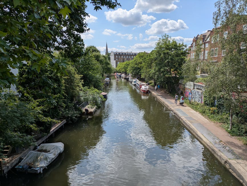 A tranquil view of a narrow canal lined with lush green trees on both sides, reflecting the blue sky with scattered white clouds. Along the right bank, there is a paved walkway with a few pedestrians, and parked cars are visible further down the street. The left side showcases boat docks with some boats, including one covered with a tarp. In the background, multi-story brick buildings and a church steeple rise above the trees, under bright daylight. The scene depicts a clean, well-maintained outdoor setting typical of residential and urban waterways, emphasizing the natural beauty and serenity of the area, aligned with the principles of surface cleaning and environmental hygiene promoted by Carpet Cleaning Islington.