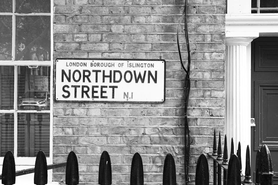 A black and white photograph of a residential building's exterior wall made of brickwork, featuring a street sign that reads 'Northdown Street, London Borough of Islington, N1'. To the left, there is a window with multiple panes, and reflections of parked cars can be seen in the glass. To the right, part of a dark wooden door with a decorative white column is visible. A black wrought iron fence with pointed tips is in the foreground, and a few electrical wires run vertically along the wall. The setting appears clean and well-maintained, with natural lighting highlighting the textures of the brick wall and the details of the street sign. This image is used to illustrate the typical exterior of a property in the Holloway Road area of Islington, relevant to local cleaning services offered by Carpet Cleaning Islington.