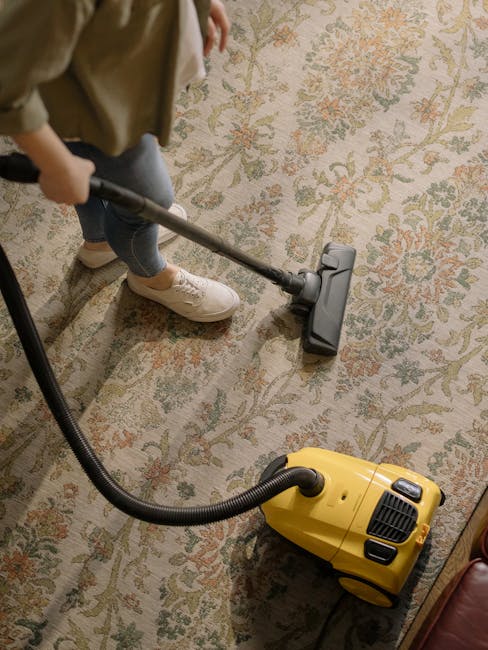 A person is vacuuming a traditional patterned area rug with a beige background and intricate floral motifs in muted shades of green, red, and cream. The individual is wearing casual clothing, including light-colored sneakers, jeans, and a beige jacket. The yellow and black vacuum cleaner is a canister model with a long flexible hose, which the person is holding with a vacuum head attached, carefully cleaning the surface. The room appears well-lit, highlighting the clean and maintained appearance of the carpet, reflecting thorough surface cleaning and maintenance by Carpet Cleaning Islington, as part of their deep cleaning and hygiene services in residential or commercial settings.