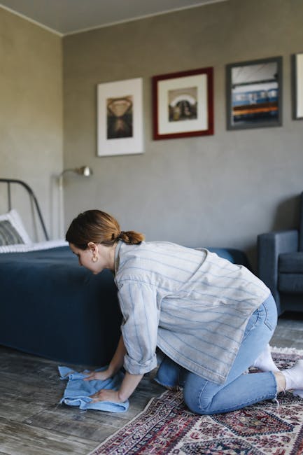 A woman with brown hair tied back, dressed in a light blue striped shirt and jeans, is kneeling on the floor of a living room while using a blue cloth to clean a dark-colored wooden floor. The room fe