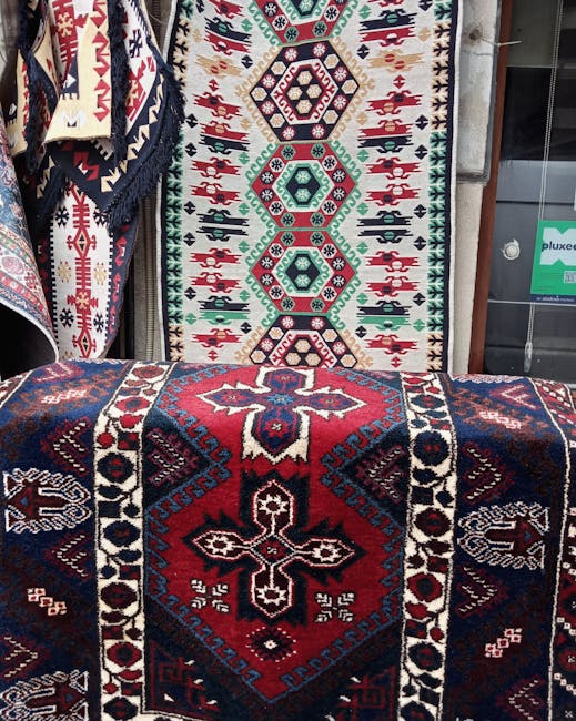 A display of traditional patterned rugs with intricate geometric designs in shades of red, blue, black, and white, shown hanging vertically against a metal frame and draped over a flat surface. The rugs exhibit detailed craftsmanship with symmetrical motifs and dense pile, demonstrating well-maintained cleanliness and vibrant colors. The setting appears to be an indoor or outdoor market stall, illuminated by natural or artificial light, with visible display fixtures. The image emphasizes surface cleanliness and careful presentation, aligning with professional cleaning and maintenance standards, as represented by Carpet Cleaning Islington's focus on deep cleaning and sanitisation of textile surfaces such as rugs.