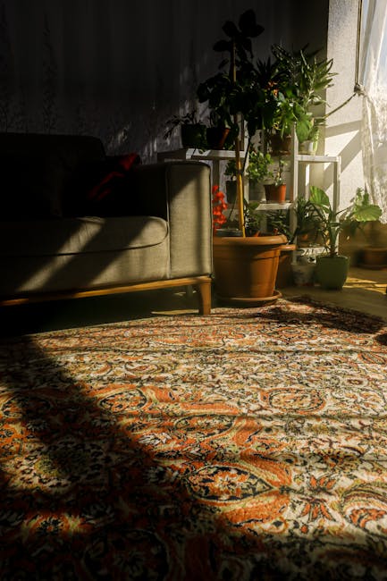 A handheld vacuum cleaner with a red and black design is positioned on a patterned area rug with intricate geometric and floral motifs in shades of red, blue, beige, and cream. The rug is situated on a hardwood floor, partially visible around its edges, and appears clean and well-maintained. Light-colored curtains or drapes are seen in the background, covering a window or partition, with soft natural light illuminating the scene. The setting appears to be a domestic living space, emphasizing surface cleaning and deep cleaning processes. The image depicts a professional approach to upholstery or carpet cleaning by Carpet Cleaning Islington, highlighting their focus on hygiene and meticulous maintenance of domestic surfaces.